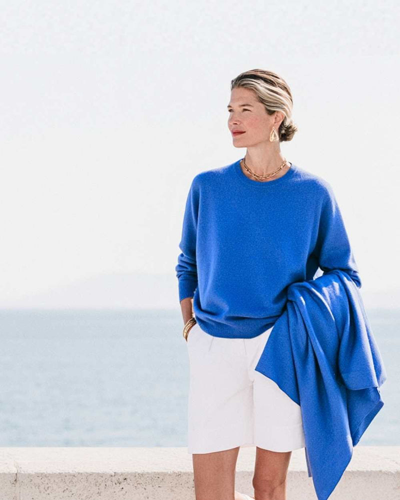 A woman on holidays by the sea wearing a bright blue knit cashmere sweater with a matching azure blue cashmere travel scarf draped over one arm.