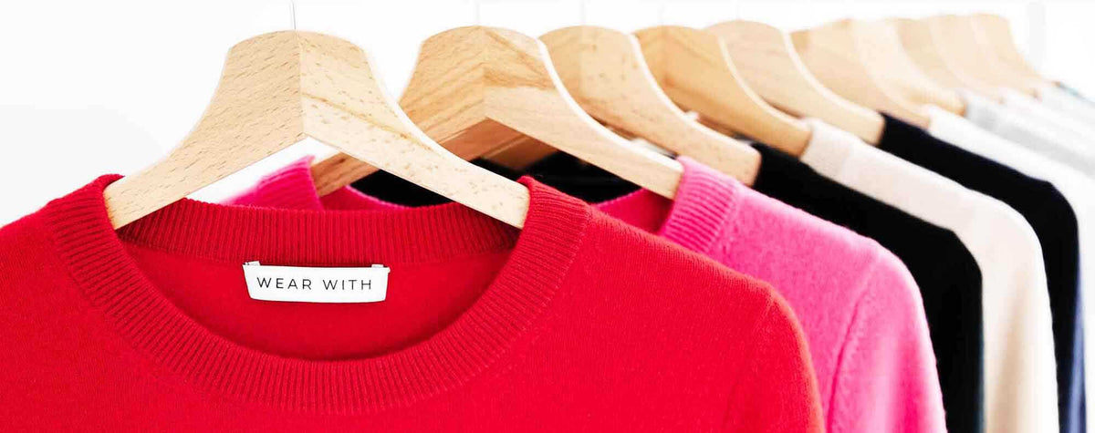 A close-up row of wooden hangers holding knit crewneck sweaters in red, pink, black, white, and light wood tones. A small neck label on the front sweater reads 'WEAR WITH'.
