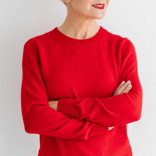 A woman wearing a bright red crewneck sweater, shown from the chest up with her arms crossed, against a plain light gray background.